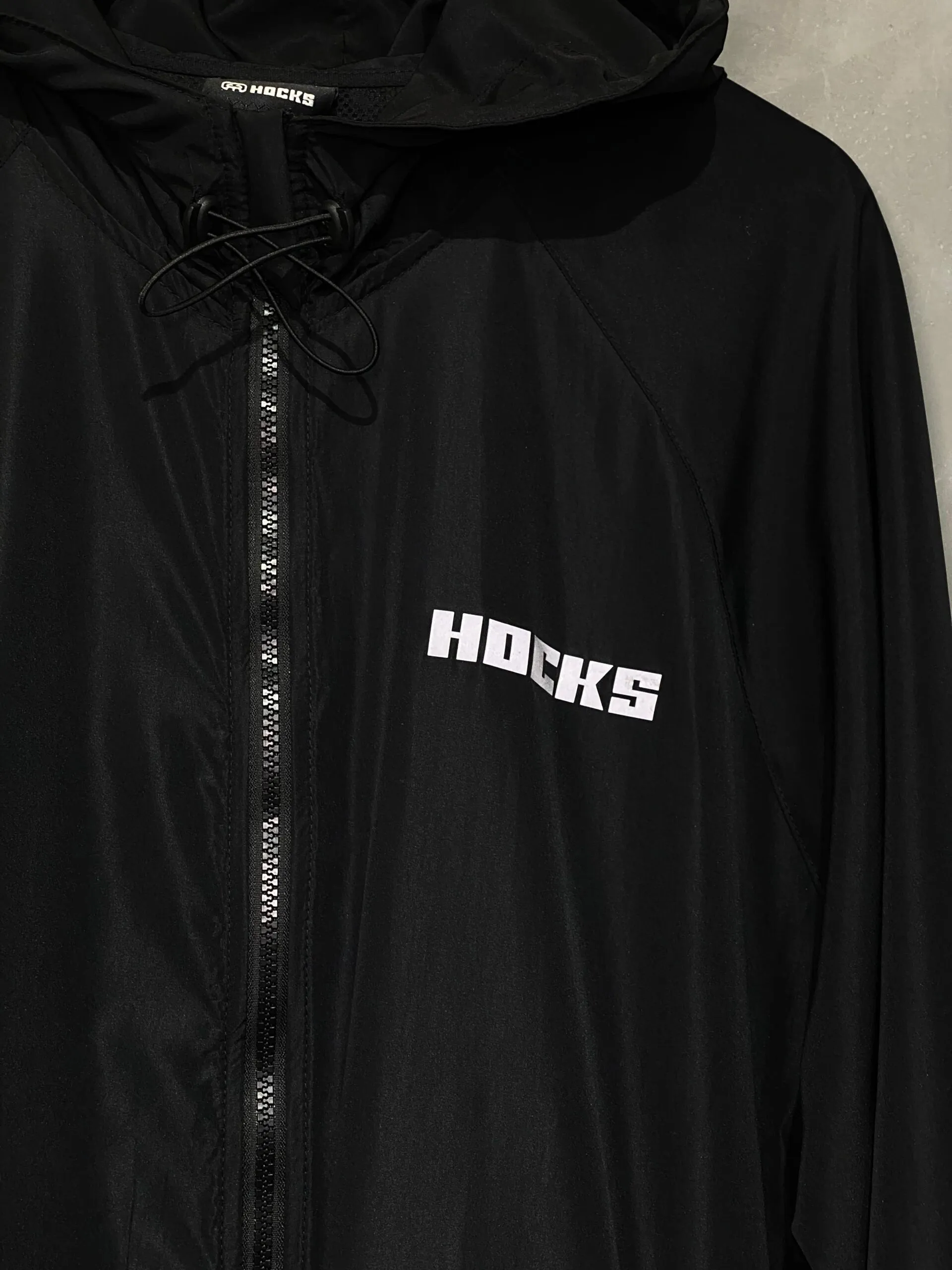 Jaqueta corta vento Hocks com logo refletivo na cor preta, ideal para proteção e estilo urbano.