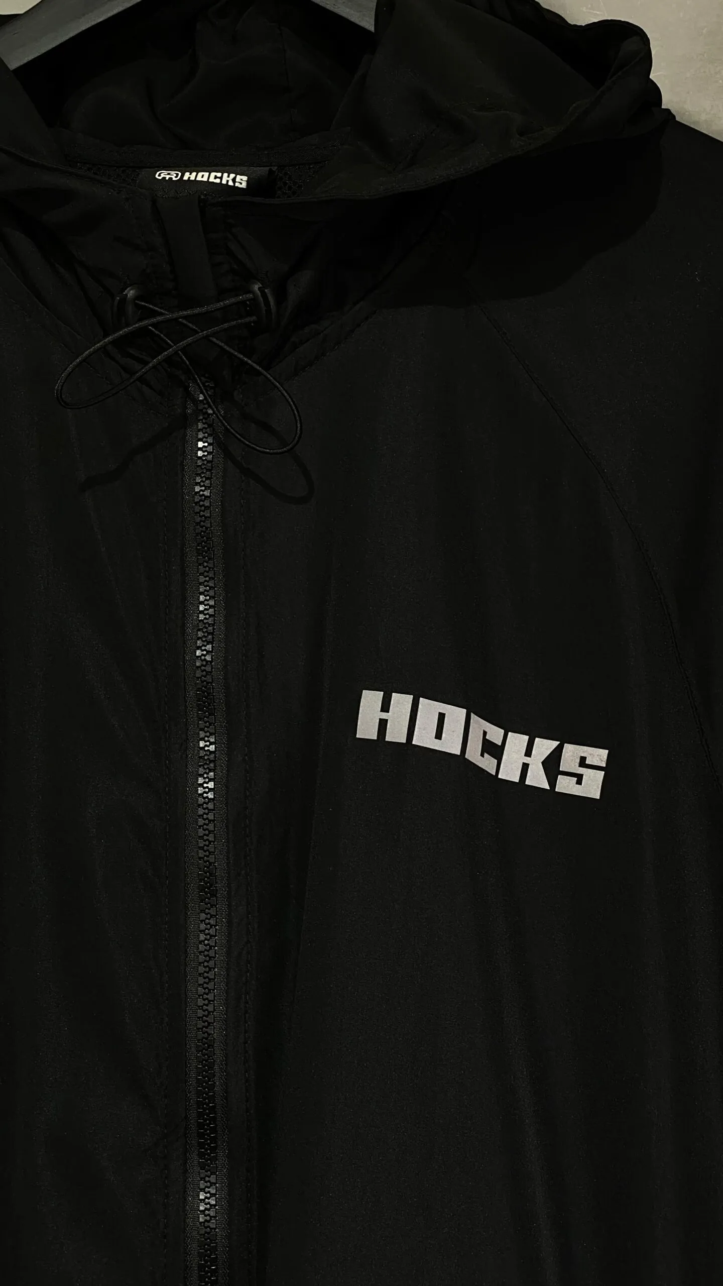 Jaqueta corta vento Hocks com logo refletivo na cor preta, ideal para proteção e estilo urbano.